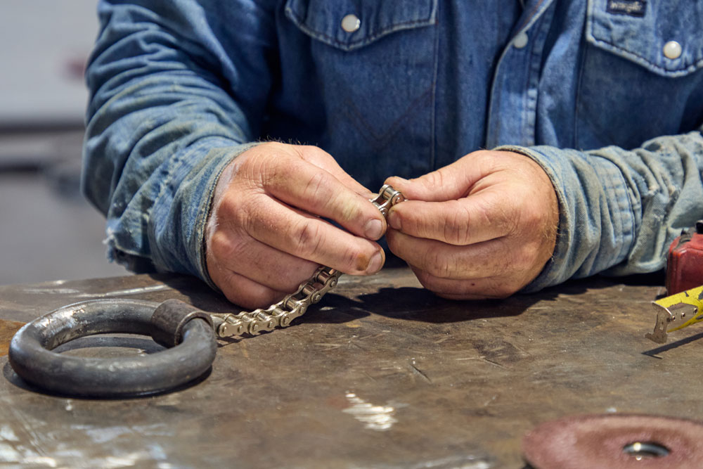 Primer plano de un hombre con camisa vaquera trabajando con una cadena metálica en una mesa de taller, rodeado de herramientas como una cinta métrica y un grillete.